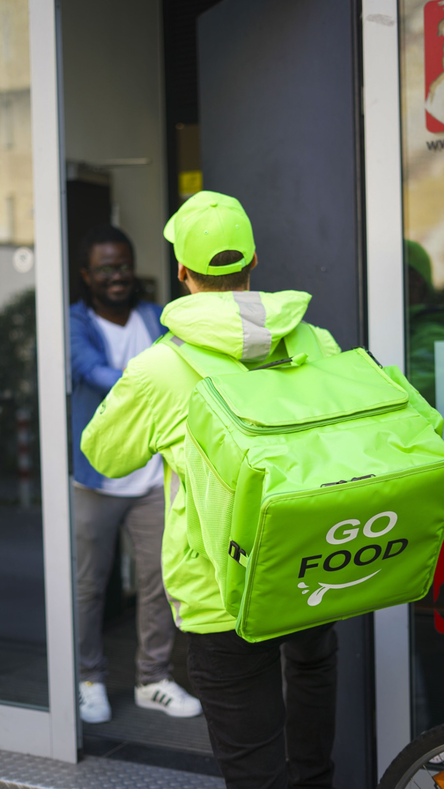 Delivery worker in ‘GO FOOD’ uniform standing at a glass door, ready to enter or exit, with another individual visible inside.
