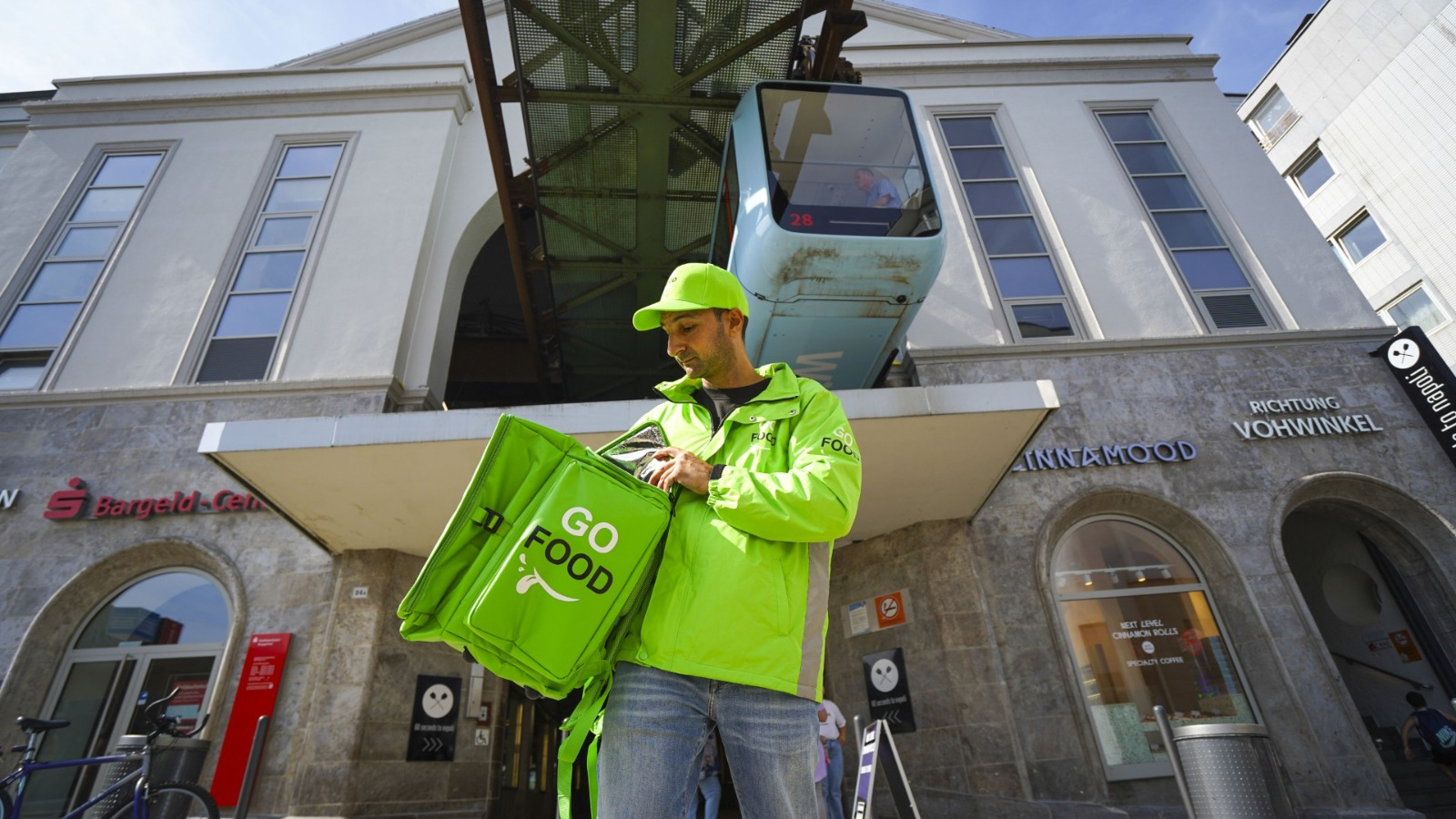 Food delivery worker in green attire ready for service in an urban setting with a tram passing by in the background.
