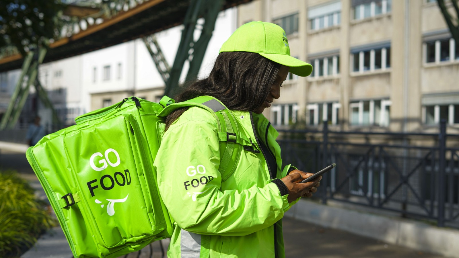 Delivery worker in ‘GO FOOD’ uniform using a smartphone, with a bridge and city buildings in the background