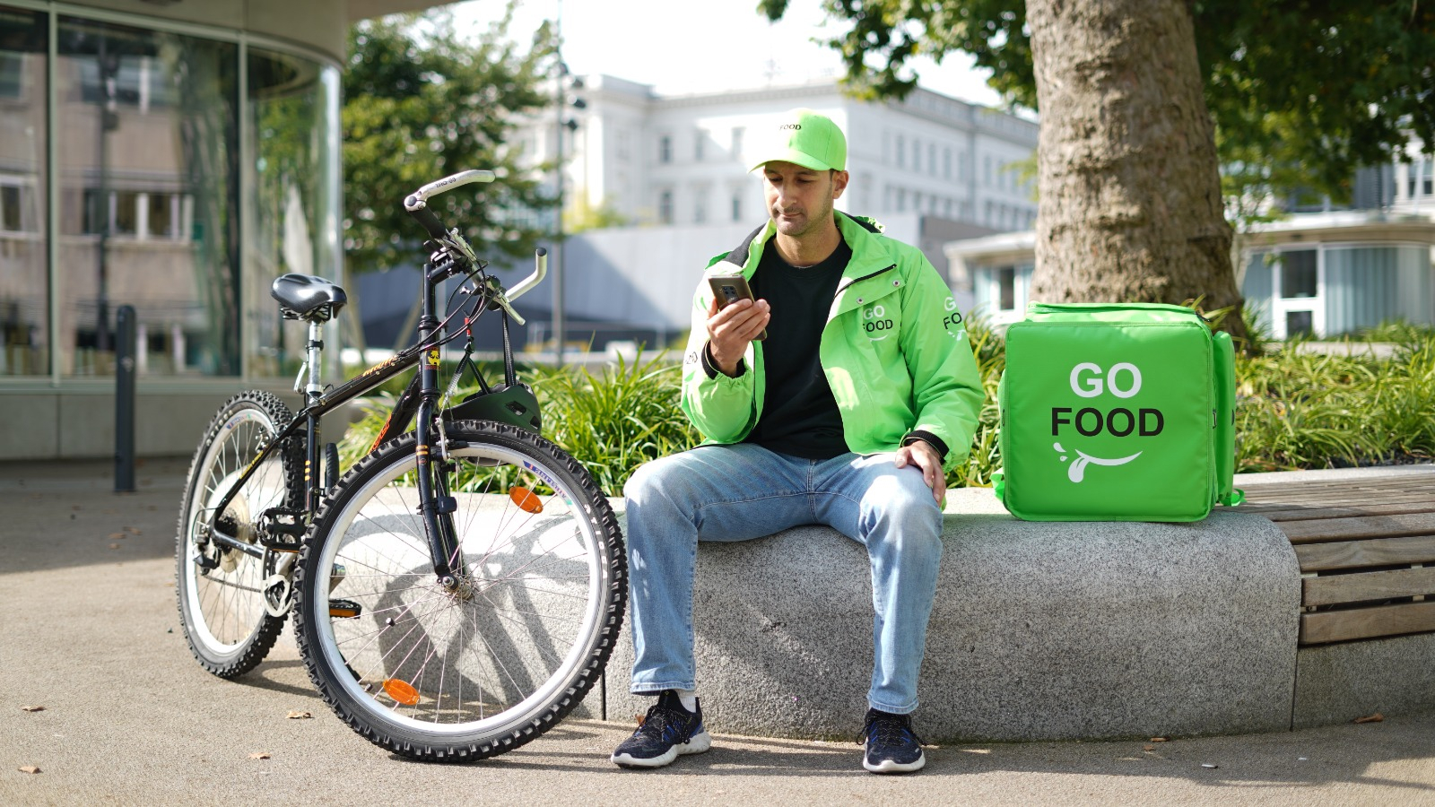 Food delivery worker in ‘GO FOOD’ attire taking a break on a bench with their bicycle and delivery bag nearby, amidst an urban backdrop.