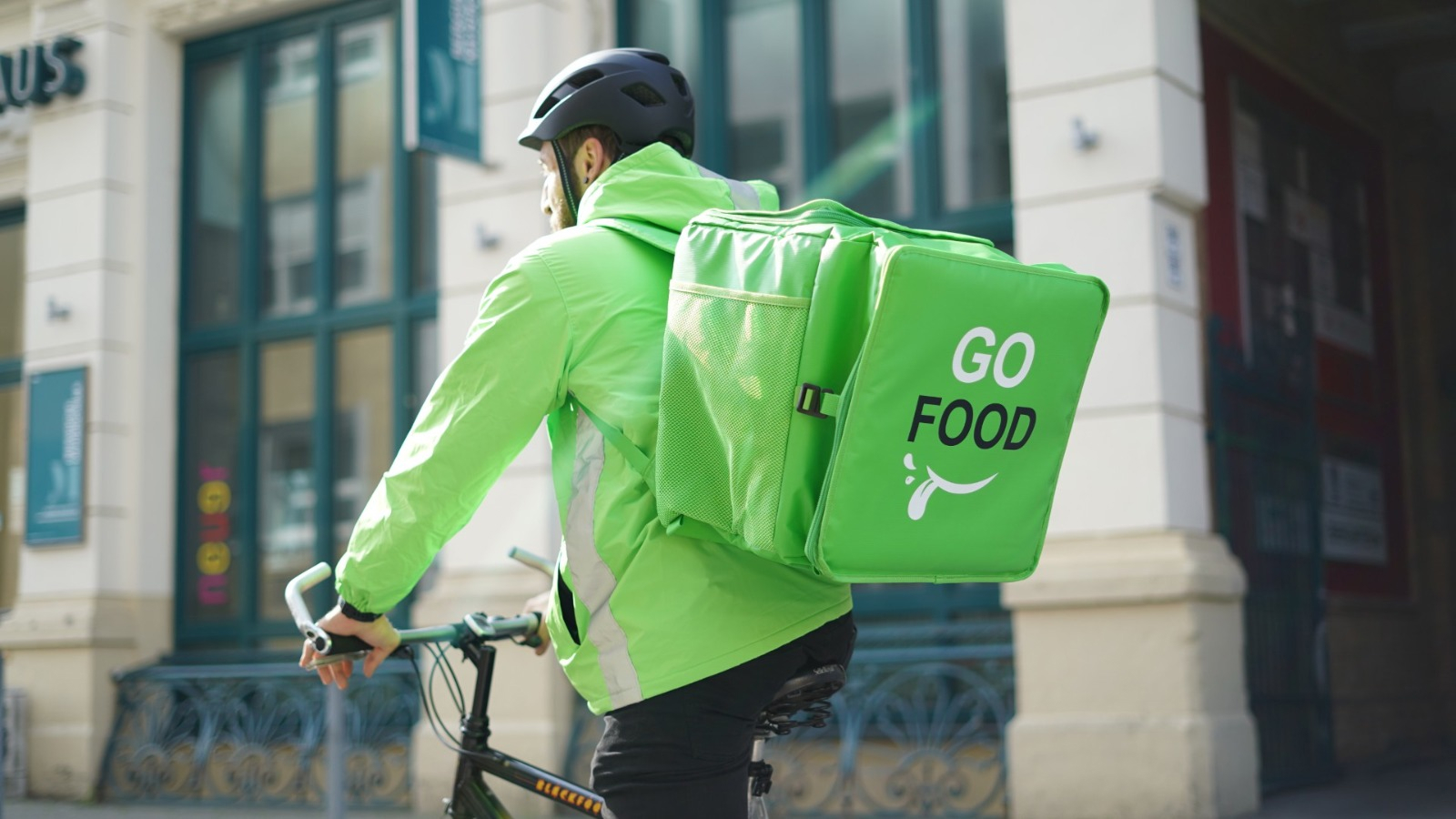 Delivery person on a bicycle with a ‘GO FOOD’ insulated backpack, navigating through an urban street.