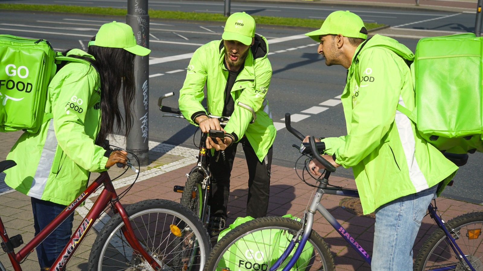 Three GO FOOD delivery workers with bicycles at a street corner, one using a mobile device, likely coordinating their next delivery.