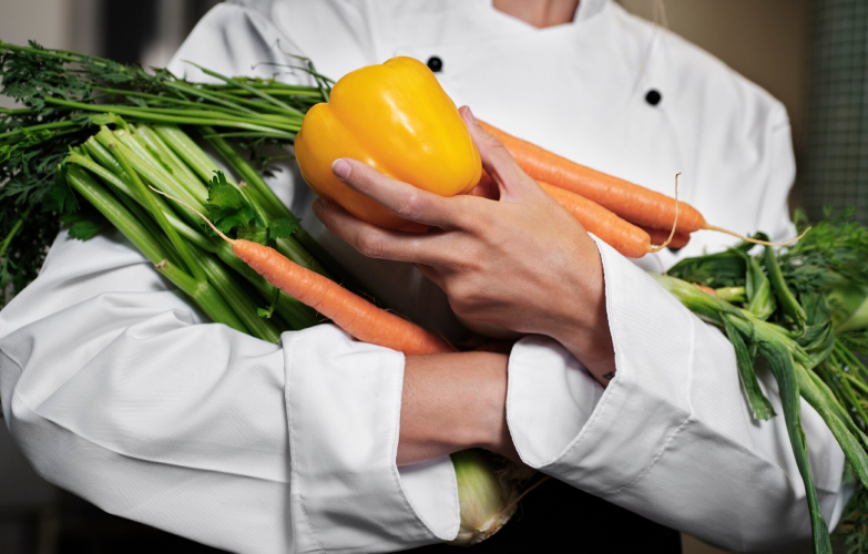 Professional chef holding carrots, lettuce, and other vegetables while cooking in a commercial kitchen.